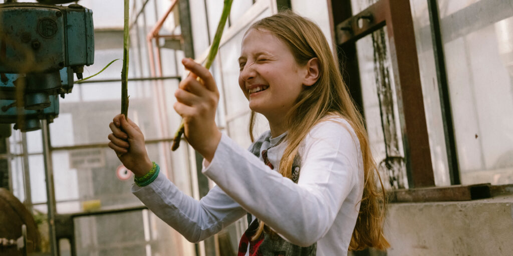 Smiling girl in an old greenhouse, caught mid-drum, beating with two drumsticks in her hands.